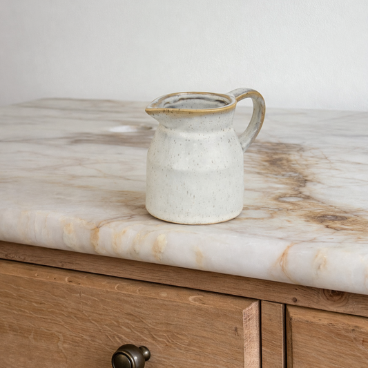 White ceramic pitcher with gold rim on a marble countertop next to wooden cabinets.