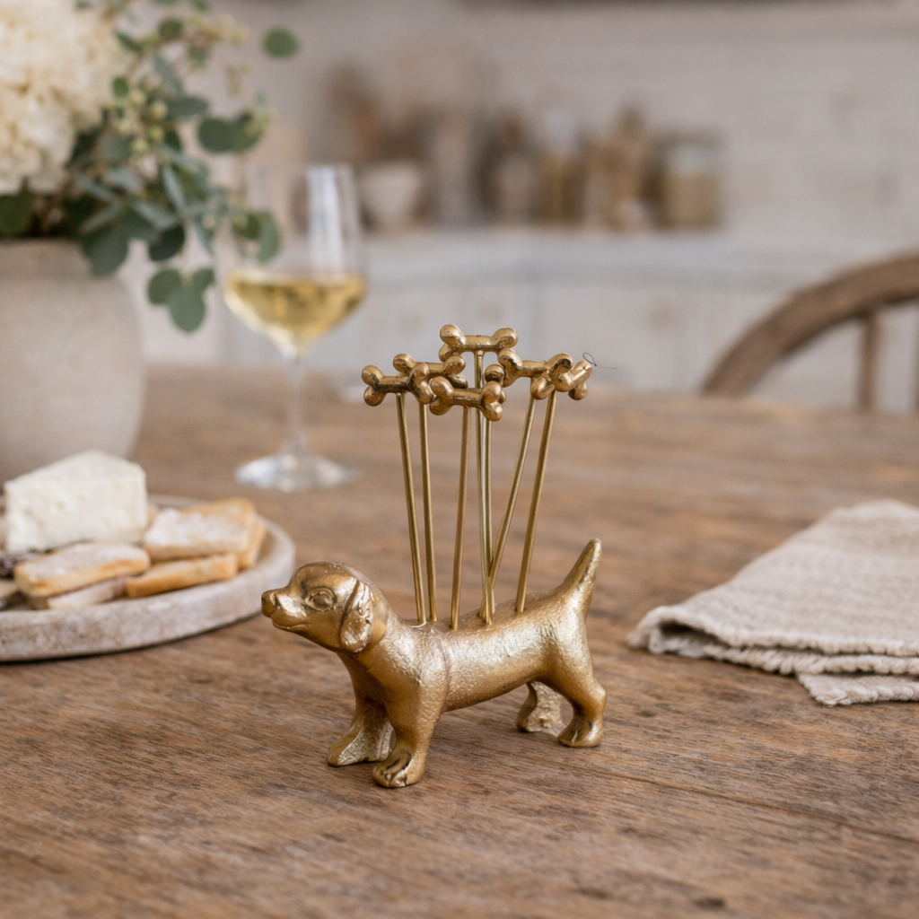 Gold dog-shaped toothpick holder on a wooden table with a glass of wine and snacks in the background.