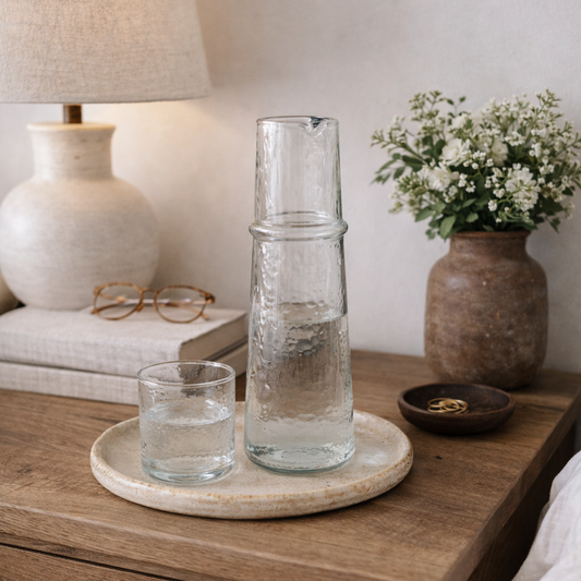 Wooden nightstand with glass vase, candle, and lamp in a cozy bedroom setting